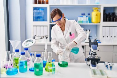 Middle age woman wearing scientist uniform pouring liquid at laboratory