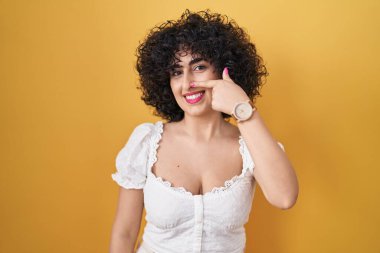 Sarı arka planda duran kıvırcık saçlı genç esmer kadın el ve yüz parmaklarını ve burnunu göstererek neşeli bir şekilde gülümsüyor. güzellik konsepti 