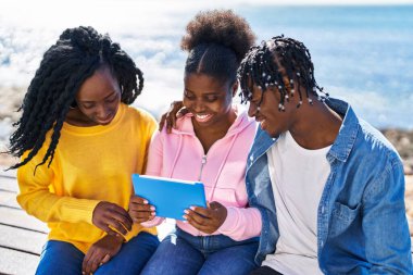 African american friends watching video on touchpad sitting on bench at seaside