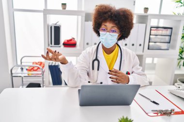 Doktor üniforması giyen ve sağlık maskesi takan Afrikalı Amerikalı kadın klinikte video görüşmesi yapıyor.