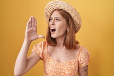 Young redhead woman standing over yellow background wearing summer hat shouting and screaming loud to side with hand on mouth. communication concept. 