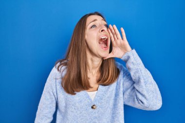 Young woman standing over blue background shouting and screaming loud to side with hand on mouth. communication concept. 