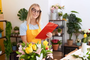 Young blonde woman florist reading document touching plant at flower shop