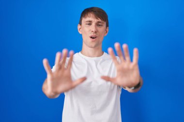 Caucasian blond man standing over blue background doing stop gesture with hands palms, angry and frustration expression 