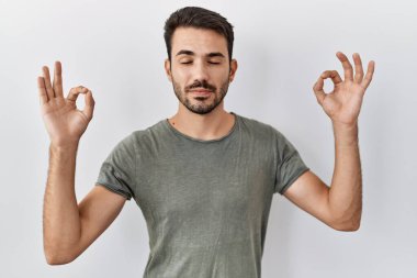 Young hispanic man with beard wearing casual t shirt over white background relax and smiling with eyes closed doing meditation gesture with fingers. yoga concept. 