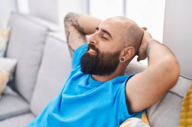 Genç kel adam, ellerini başının üstüne koyup evinde kanepede oturuyor.