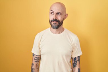 Sarı arka planda duran İspanyol dövmeli adam gülümseyip yan tarafa bakıyor ve düşünüyor.. 