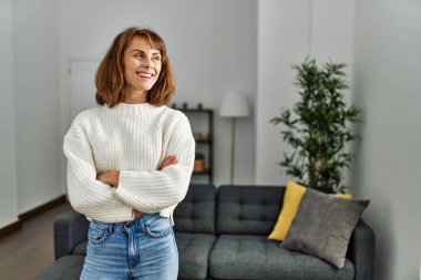 Beyaz bir kadın gülümsüyor. Kollarını kavuşturup evde duruyor.