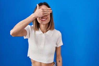 Mavi arka planda dikilen İspanyol kadın. Gülümsüyor ve gülümsüyor. Gözlerini şaşkınlıkla kapatıyor. kör kavram. 