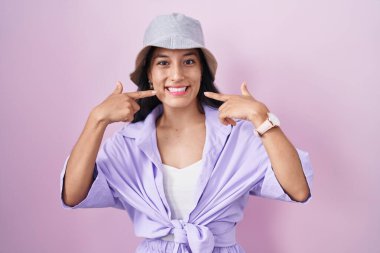 Young hispanic woman standing over pink background wearing hat smiling cheerful showing and pointing with fingers teeth and mouth. dental health concept. 