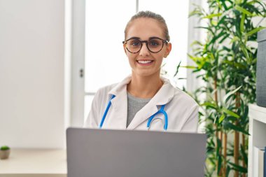 Young blonde woman wearing doctor uniform using laptop working at clinic