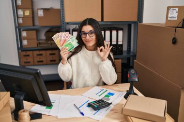 Young hispanic woman working at small business ecommerce holding money doing ok sign with fingers, smiling friendly gesturing excellent symbol 