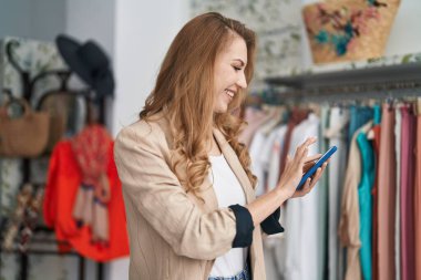 Giyim mağazasında akıllı telefon kullanarak gülümseyen genç sarışın kadın müşteri.