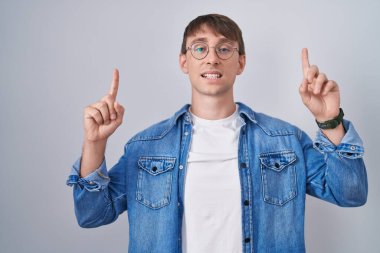Caucasian blond man standing wearing glasses smiling amazed and surprised and pointing up with fingers and raised arms. 