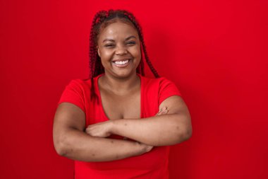African american woman with braided hair standing over red background happy face smiling with crossed arms looking at the camera. positive person. 