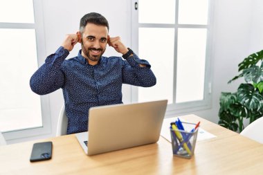 Ofiste çalışan sakallı, laptopuyla gülümseyen kulakları parmaklarıyla çeken, komik hareketler yapan genç bir İspanyol. deneme sorunu 