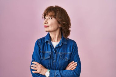 Orta yaşlı bir kadın pembe arka planda duruyor. Kollarını kavuşturmuş, ikna olmuş ve kendinden emin bir şekilde yan tarafa bakıyor. 