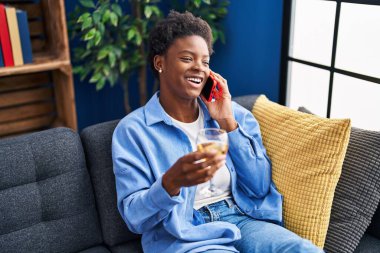 Afro-Amerikalı kadın akıllı telefondan konuşuyor evde şampanya içiyor.