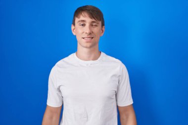 Caucasian blond man standing over blue background winking looking at the camera with sexy expression, cheerful and happy face. 