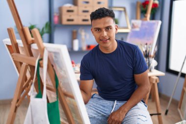 Young hispanic man painting sitting at art studio looking positive and happy standing and smiling with a confident smile showing teeth 