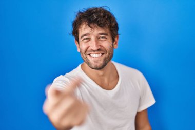 Hispanik genç adam mavi arka planda duruyor. Buraya el kol hareketi yaparak mutlu ve gülümseyen insanları davet ediyor. 