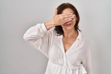 Orta yaşlı İspanyol bir kadın izole edilmiş arka planda duruyor. Gülümsüyor ve gülümsüyor. Gözlerini şaşkınlıkla kapatıyor. kör kavram. 