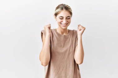 İzole edilmiş arka planda duran genç sarışın kadın başarı için heyecanlanıyor kolları havada ve gözleri kapalı zaferi kutlarken gülümsüyor. Kazanan konsept. 