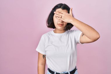 Young hispanic woman wearing casual white t shirt over pink background covering eyes with hand, looking serious and sad. sightless, hiding and rejection concept 