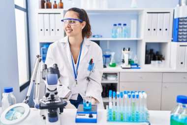 Young hispanic woman working at scientist laboratory looking away to side with smile on face, natural expression. laughing confident. 