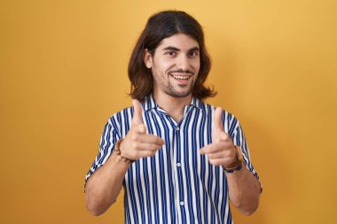 Hispanic man with long hair standing over yellow background pointing fingers to camera with happy and funny face. good energy and vibes. 