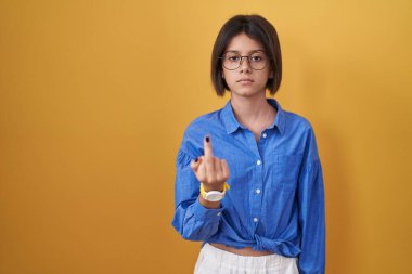 Young girl standing over yellow background showing middle finger, impolite and rude fuck off expression 