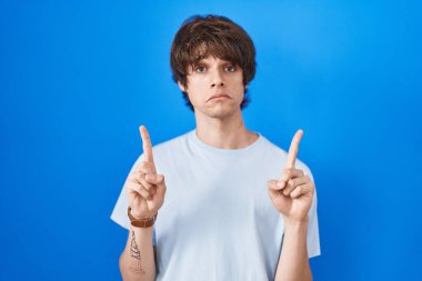 Mavi arka planda dikilen İspanyol genç adam üzgün ve üzgün görünüyor, parmaklarıyla yön gösteriyor, mutsuz ve depresif.. 