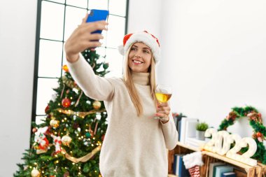 Genç sarışın kadın akıllı telefonun yanında selfie çekiyor. Noel ağacının yanında duruyor.