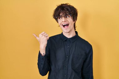 Young man wearing glasses over yellow background smiling with happy face looking and pointing to the side with thumb up. 
