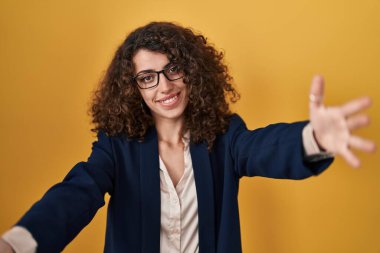 Hispanic woman with curly hair standing over yellow background looking at the camera smiling with open arms for hug. cheerful expression embracing happiness. 