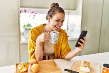 Genç ve güzel İspanyol kadın kahvaltısını yapıyor. Mutfakta akıllı telefon kullanarak kahve içiyor.