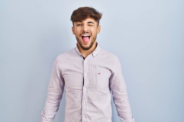 Arab man with beard standing over blue background sticking tongue out happy with funny expression. emotion concept. 
