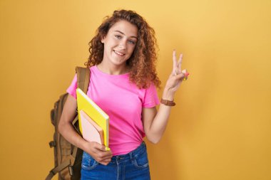 Sırt çantası takan, elinde kitaplarla kameraya bakıp zafer işareti yapan beyaz bir kadın. İki numara.. 
