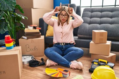 Young hispanic woman moving to a new home sitting on the floor doing funny gesture with finger over head as bull horns 