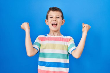Mavi arka planda dikilmiş genç beyaz çocuk kolları havada ve gözleri kapalı bir şekilde başarıyı kutluyor ve şaşırıyor. Kazanan konsept. 