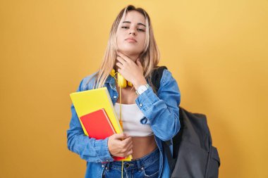 Young blonde woman wearing student backpack and holding books touching painful neck, sore throat for flu, clod and infection 