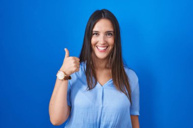 Mavi arka planda duran genç esmer kadın elleriyle mutlu bir jest yapıyor. Kameraya bakıp başarıyı gösteren ifade. 