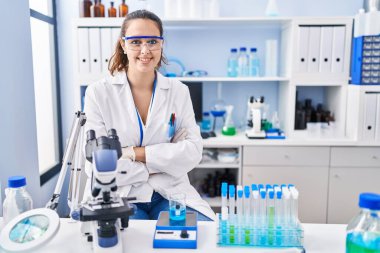 Young hispanic woman working at scientist laboratory happy face smiling with crossed arms looking at the camera. positive person. 