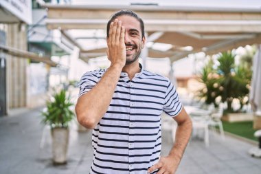 Young hispanic man with beard outdoors at the city covering one eye with hand, confident smile on face and surprise emotion. 