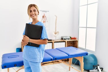 Genç beyaz fizyoterapist klinikte mutlu bir şekilde klasörü tutuyor.