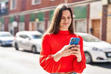 Genç, güzel, İspanyol bir kadın sokakta akıllı telefon kullanarak gülümsüyor.