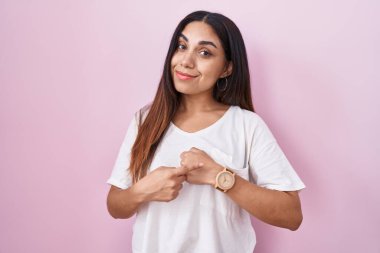 Young arab woman standing over pink background in hurry pointing to watch time, impatience, looking at the camera with relaxed expression 
