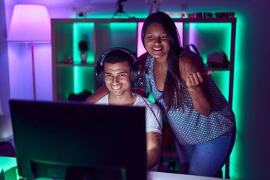 Genç İspanyol çift video oyunları oynuyor başparmağıyla yan tarafa işaret ediyor ve ağzı açık gülümsüyor. 