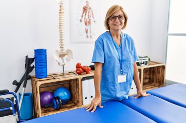 Klinikte fizyoterapist üniforması giyen orta yaşlı sarışın kadın.