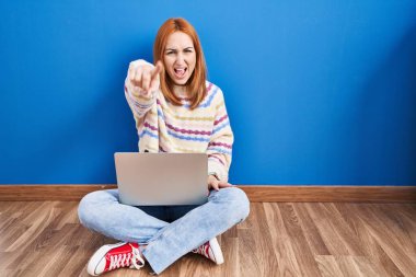 Evde dizüstü bilgisayar kullanan genç bir kadın yerde oturmuş kameraya kızgın ve sinirli bir şekilde seni işaret ediyor. 